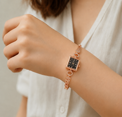A close-up of a woman's wrist wearing a rose gold watch with a black dial and a delicate chain bracelet.
