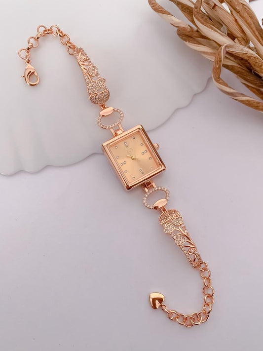 A rose gold women's watch with a square face and ornate chain bracelet, styled on a light gray surface with a white scalloped dish and dried grass in the corner.