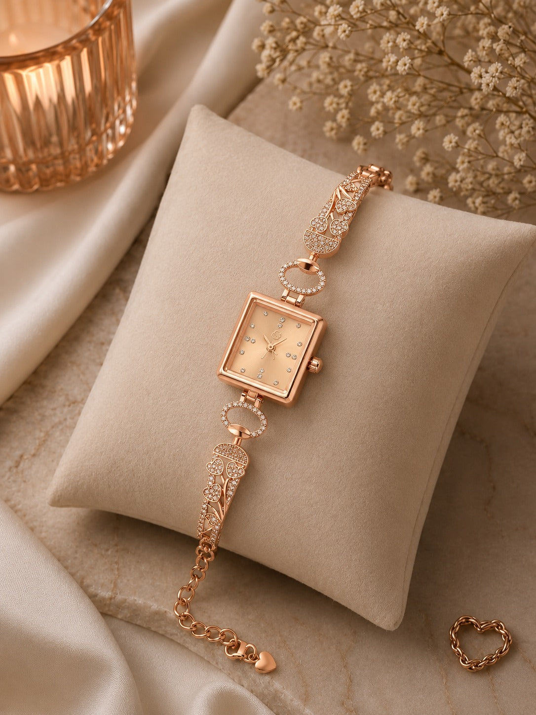 A rose gold women's watch with a square dial and wing-shaped links, displayed on a beige fabric cushion with decorative satin, dried flowers, and a candle.
