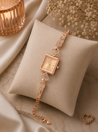 A rose gold women's watch with a square dial and wing-shaped links, displayed on a beige fabric cushion with decorative satin, dried flowers, and a candle.