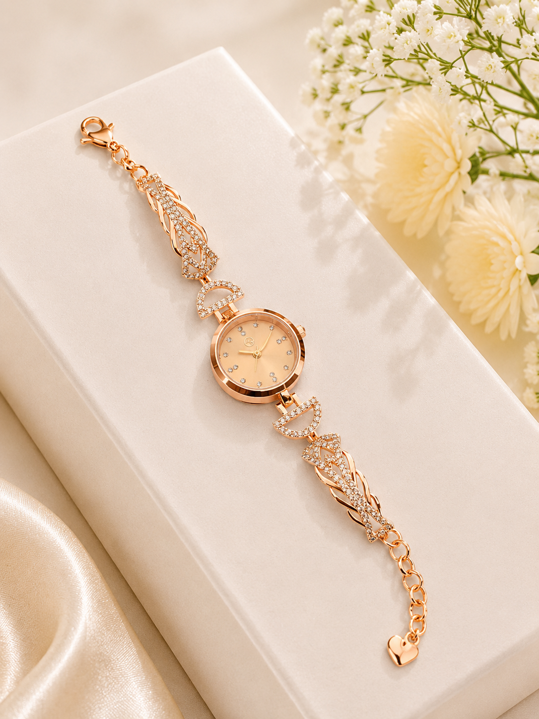 A rose gold women's watch with a delicate link bracelet adorned with sparkling clear crystals, resting on a white gift box with soft cream flowers in the background.