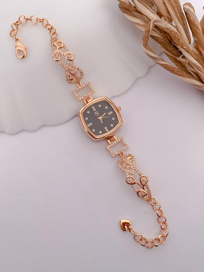 A rose gold women's watch with a square face and a decorative chain bracelet featuring bow and heart motifs, displayed on a white surface with a dried floral prop.
