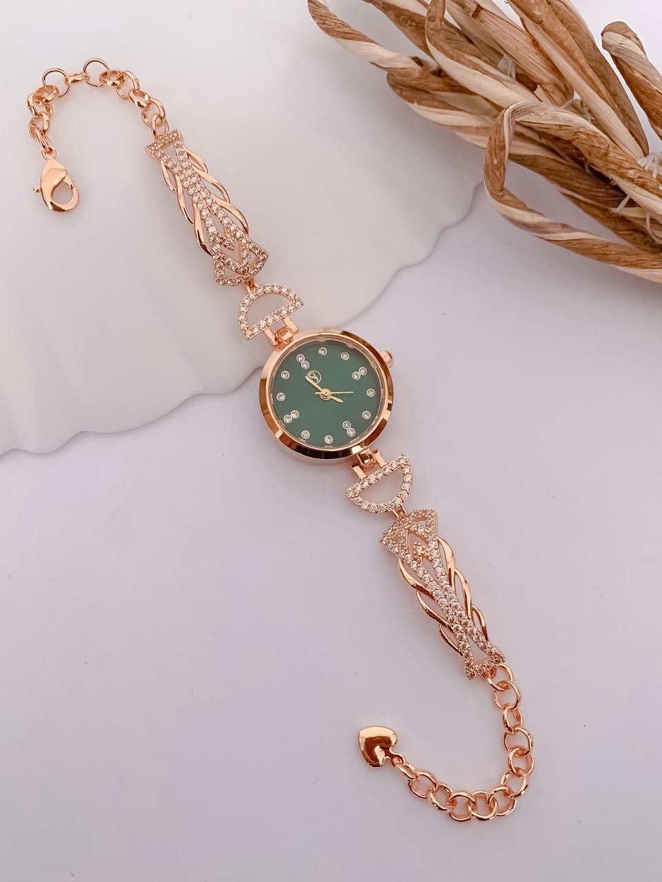 A rose gold watch with a round green dial and crystal hour markers, featuring an ornate link bracelet with crystal accents, displayed on a white background with dried grass.