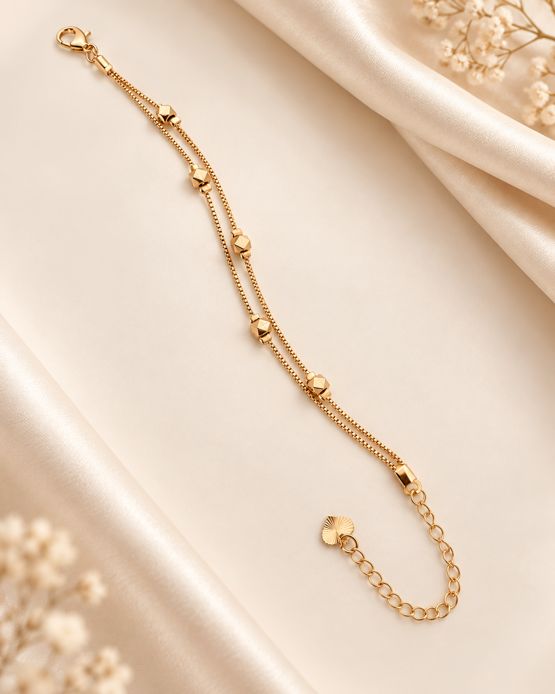 A gold-toned double-strand chain anklet with small heart-shaped beads and a lobster clasp, displayed on a cream satin fabric background with dried floral accents.