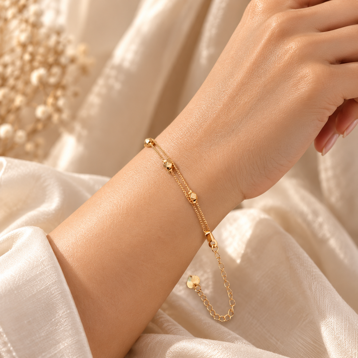 A delicate gold chain bracelet with small bead details and a charm, shown worn on a person's wrist against a soft cream-colored fabric background.