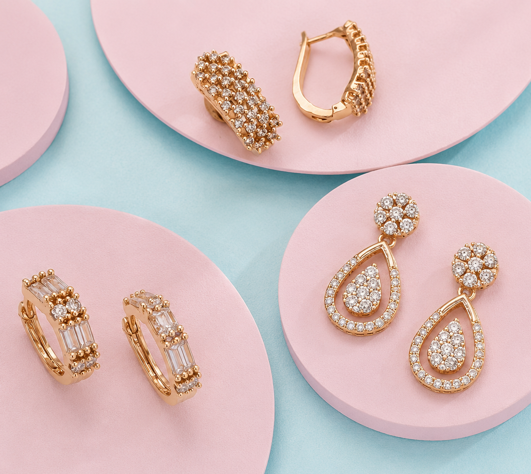 Four pairs of gold-toned jewelry displayed on pink circular platforms against a light blue background: two hoop-style earrings, two teardrop drop earrings with pavé and clear stones.